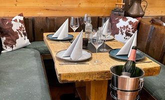A cozy table in a rustic restaurant, decorated with folded napkins and glasses. A bottle of champagne is ready in a bucket. | © Kuhstall | Uwe Nagel
