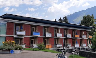 A modern building with several floors and balconies. In the background, green hills and a blue sky can be seen. | © Kleinwalsertal Tourismus | Veronika Senn