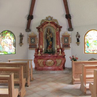 A small chapel with wooden bench rows and colorful windows. In the foreground stands an ornate altar with a statue. | © Stefan Heim