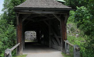 Eine Holzbrücke führt durch einen überdachten Gang. Umgeben von grünen Bäumen und einem klaren Himmel wirkt die Umgebung friedlich. | © Stefan Heim