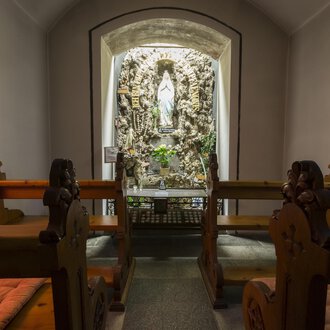 A quiet chapel with wooden benches and a statue in the niche. The walls are softly illuminated, creating a peaceful atmosphere. | © Kleinwalsertal Tourismus | Oliver Farys