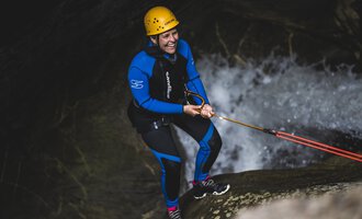 A person in a blue wetsuit and a helmet stands at a waterfall. They are pulling with a rope and have a broad smile on their face. | © MAP-Erlebnis
