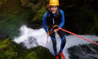 An adventurous person rappelling down a small waterfall. They are wearing a helmet and a wetsuit and smiling at the camera. | © MAP-Erlebnis