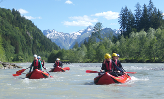 Four kayakers are paddling on a calm river, surrounded by green mountains and snow-capped peaks. It is a sunny day with a blue sky and a few clouds. | © MAP-Erlebnis