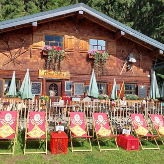 A traditional wooden house surrounded by trees. In front of the house are deck chairs and umbrellas in a friendly atmosphere. | © Maxhütte | Viera Krafcik
