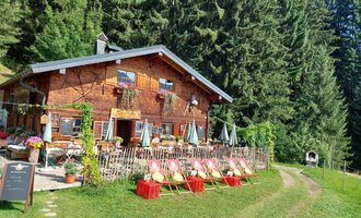 Ein gemütliches Holzhaus umgeben von hohen Bäumen. Vor dem Haus stehen Stühle und Tische in einer einladenden Außenanlage. | © Maxhütte | Viera Krafcik