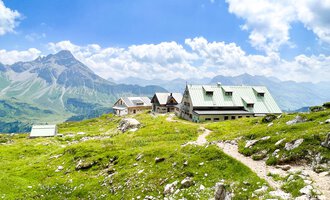 A picturesque mountain landscape with green grass and a clear sky. In the foreground, typical mountain chalets can be seen. | © Mindelheimer Hütte | Wolfgang Stache