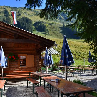 Eine gemütliche Hütte in den Bergen mit Tischen und Bänken im Freien. Im Hintergrund sind grüne Wiesen und Berge zu sehen. | © Kleinwalsertal Tourismus