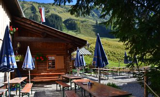 Eine gemütliche Hütte in den Bergen mit Tischen und Bänken im Freien. Im Hintergrund sind grüne Wiesen und Berge zu sehen. | © Kleinwalsertal Tourismus