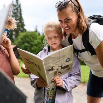 A mother is reading with her children in nature. The children are looking curiously at a book. | © Kleinwalsertal Tourismus | Oliver Farys
