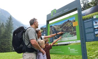 A family is standing in front of an information board in nature. They are pointing at the map and discussing the surroundings. | © Kleinwalsertal Tourismus | Oliver Farys