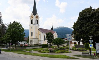 A quiet street in a charming village with a tall church in the background. Surrounded by trees and mountains, the scene radiates tranquility. | © Kleinwalsertal Tourismus | Oliver Farys