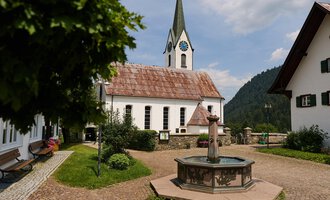 Ein malerisches Dorf mit traditionellen Häusern und einer Kirche im Hintergrund. In der Mitte befindet sich ein Brunnen, umgeben von grünem Gras und Bäumen. | © Kleinwalsertal Tourismus | Oliver Farys