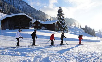 Eine Gruppe von Skifahrern fährt auf einer verschneiten Piste. Im Hintergrund sind holzige Hütten und verschneite Berge zu sehen. | © Andy Herr | Werbewind GmbH