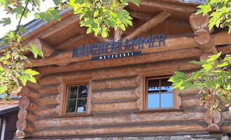 Ein rustikales Holzgebäude mit einem Schild, das "Mühlenkammer" zeigt. Umgeben von grünen Blättern und einem klaren Himmel. | © Kleinwalsertal Tourismus | Veronika Senn