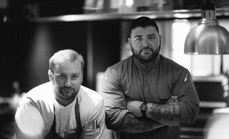 Two chefs are standing in a modern kitchen. They are looking directly at the camera and appear focused and professional.