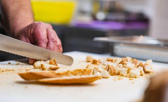 A hand is cutting bread into small cubes on a cutting board. In the background, kitchen utensils and a bowl are visible.