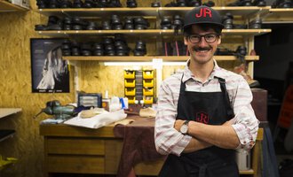 Ein Schuhmacher steht in seiner Werkstatt mit einem breiten Lächeln. Im Hintergrund sind viele Schuhe und Werkzeuge sichtbar. | © Kleinwalsertal Tourismus | Oliver Farys