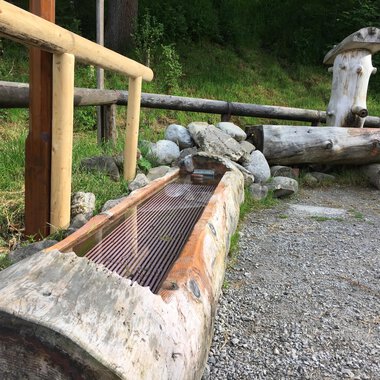 A rustic wooden fountain made from tree trunks in a natural setting. In the background, more wooden elements and vegetation can be seen. | © Kleinwalsertal Tourismus