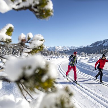 Zwei Personen skifahren auf einer verschneiten Strecke. Im Hintergrund sind schneebedeckte Berge und ein klarer blauer Himmel zu sehen. | © Kleinwalsertal Tourismus | Dominik Berchtold