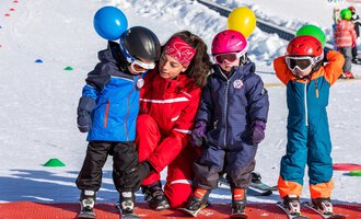Eine Skilehrerin steht zwischen vier Kindern in bunter Winterkleidung auf einem Übungshang. Die Kinder tragen Helme und haben Luftballons in den Händen. | © Skischule Bödmen-Baad GmbH