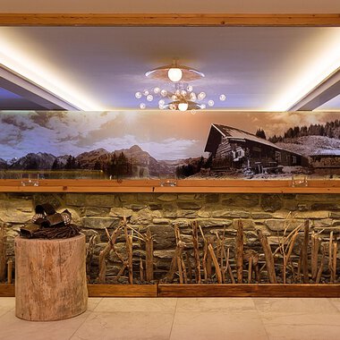 Ein elegantes Interieur mit einer Steinwand und einem großen Spiegel. Im Hintergrund ist ein großflächiges Landschaftsbild mit einer Hütte und Bergen zu sehen. | © Aparthotel Kleinwalsertal Haller GmbH & Co KG