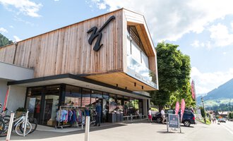 Ein modernes Geschäft mit Holzfassade und großen Fenstern. Im Vordergrund sind Kleidung und Fahrräder ausgestellt, umgeben von einer schönen Berglandschaft. | © Sport Kessler GmbH