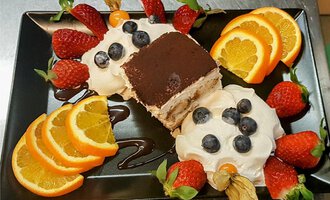 A beautiful dessert on a black plate. It consists of tiramisu, cream, and fresh fruits like strawberries and oranges.
