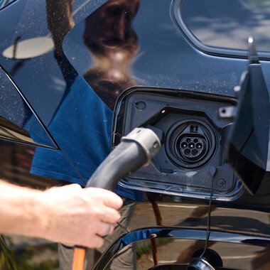 Eine Person lädt ein Elektroauto auf. Das Auto hat eine schwarze Karosserie und das Ladegerät wird angeschlossen. | © Kleinwalsertal Tourismus | Oliver Farys
