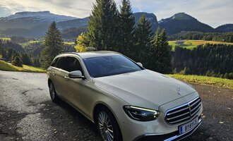 An elegant car is standing on a picturesque road in the mountains. In the background, green meadows and wooded hills can be seen. | © Taxi Beranek | Elmar Beranek