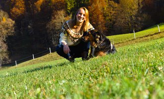 Eine Frau sitzt im Gras und lächelt neben einem Hund. Im Hintergrund sind Bäume und eine grüne Wiese zu sehen. | © Tierarztpraxis Oberstdorf | Regina Baumgartner