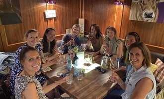 Eine Gruppe von acht Frauen sitzt an einem Tisch und hebt Gläser. Es ist eine fröhliche Atmosphäre, umgeben von Holzdekor und sanftem Licht. | © Uusziit | Chantal Kessler