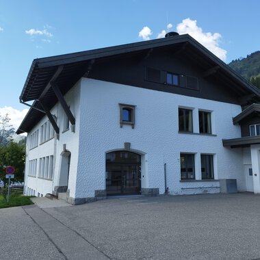 A modern, white building with a large roof. It is located in a mountainous area with green grass and trees. | © Kleinwalsertal Tourismus | N. Lughammer
