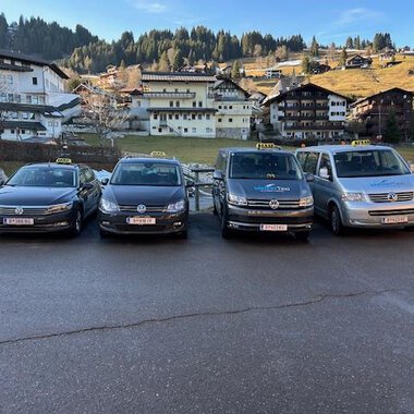 Vier Taxis stehen in einer Reihe vor einer malerischen Berglandschaft. Im Hintergrund sind traditionelle alpine Gebäude und bewaldete Hügel zu sehen. | © Walser Taxi | Josef Düringer