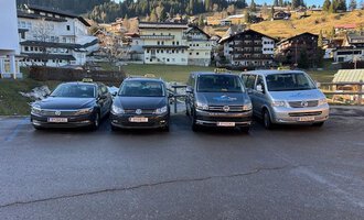 Four taxis are lined up in front of a picturesque mountain landscape. In the background, traditional alpine buildings and wooded hills can be seen. | © Walser Taxi | Josef Düringer