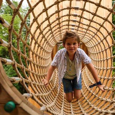 Ein Junge krabbelt durch ein großes Netz auf einem Spielplatz. Im Hintergrund sind Bäume zu sehen. | © Kleinwalsertal Tourismus | Oliver Farys