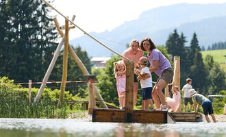 Eine Gruppe von Kindern und Erwachsenen steht auf einer Holzbrücke über einem ruhigen Teich. Im Hintergrund sind Bäume und Berge zu sehen. | © Kleinwalsertal Tourismus | Oliver Farys