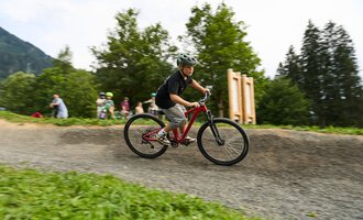 Ein Junge fährt mit einem Mountainbike auf einem geschwungenen Weg. Im Hintergrund sind weitere Kinder und Bäume zu sehen. | © Kleinwalsertal Tourismus | Oliver Farys