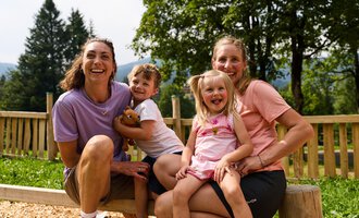 A cheerful scene with two adults and two children outdoors.  
Everyone is laughing and enjoying their time together in nature. | © Kleinwalsertal Tourismus | Oliver Farys