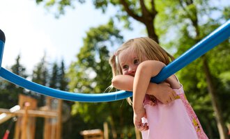 A little girl is playing in a playground. She is holding onto a blue play structure and is looking happily. | © Kleinwalsertal Tourismus | Oliver Farys