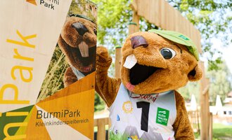 A funny mascot beaver stands next to a sign of the BurmiPark. The beaver is wearing a green hat and a sporty outfit. | © Kleinwalsertal Tourismus | Oliver Farys