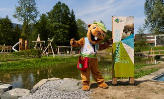 A friendly mascot in an animal costume stands next to an information sign by the water. In the background, there are playground equipment and a lot of greenery. | © Kleinwalsertal Tourismus | Oliver Farys