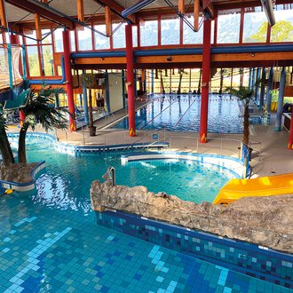 Ein farbenfrohes Hallenbad mit verschiedenen Becken und einer tropischen Atmosphäre. Palmen und Felsen schaffen ein entspannendes Ambiente. | © WONNEMAR
