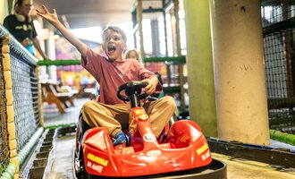 Ein fröhlicher Junge fährt in einem roten Bobby-Car und winkt begeistert. Im Hintergrund sind weitere Spielgeräte zu sehen. | © Wonniland Sonthofen | interSPA Gruppe