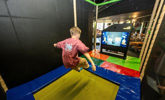 Ein Junge springt auf einem Trampolin in einem Spielbereich. Im Hintergrund ist ein Bildschirm mit Spielinhalten zu sehen. | © Wonniland Sonthofen | interSPA Gruppe