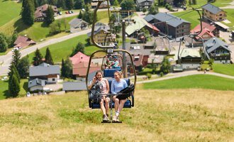 Two women are sitting in a chair lift and riding up a mountain meadow. In the background, green hills and a small village with houses can be seen. | © Kleinwalsertal Tourismus | Oliver Farys