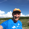 A smiling man in a blue t-shirt and orange cap outdoors in a green landscape with a blue sky and clouds. | © Kleinwalsertal Tourismus eGen
