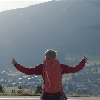 A person with a red backpack stands in a field, pointing upwards, as a helicopter flies overhead in a scenic mountainous area. | © Kleinwalsertal Tourismus eGen
