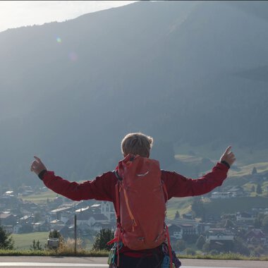 A person with a red backpack stands in a field, pointing upwards, as a helicopter flies overhead in a scenic mountainous area. | © Kleinwalsertal Tourismus eGen