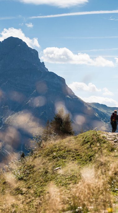 A hiker walking on a trail in the mountains of the Black Forest on a sunny day, surrounded by green grass and mountain peaks in the background. | © Kleinwalsertal Tourismus eGen • Photographer: Andre Tappe